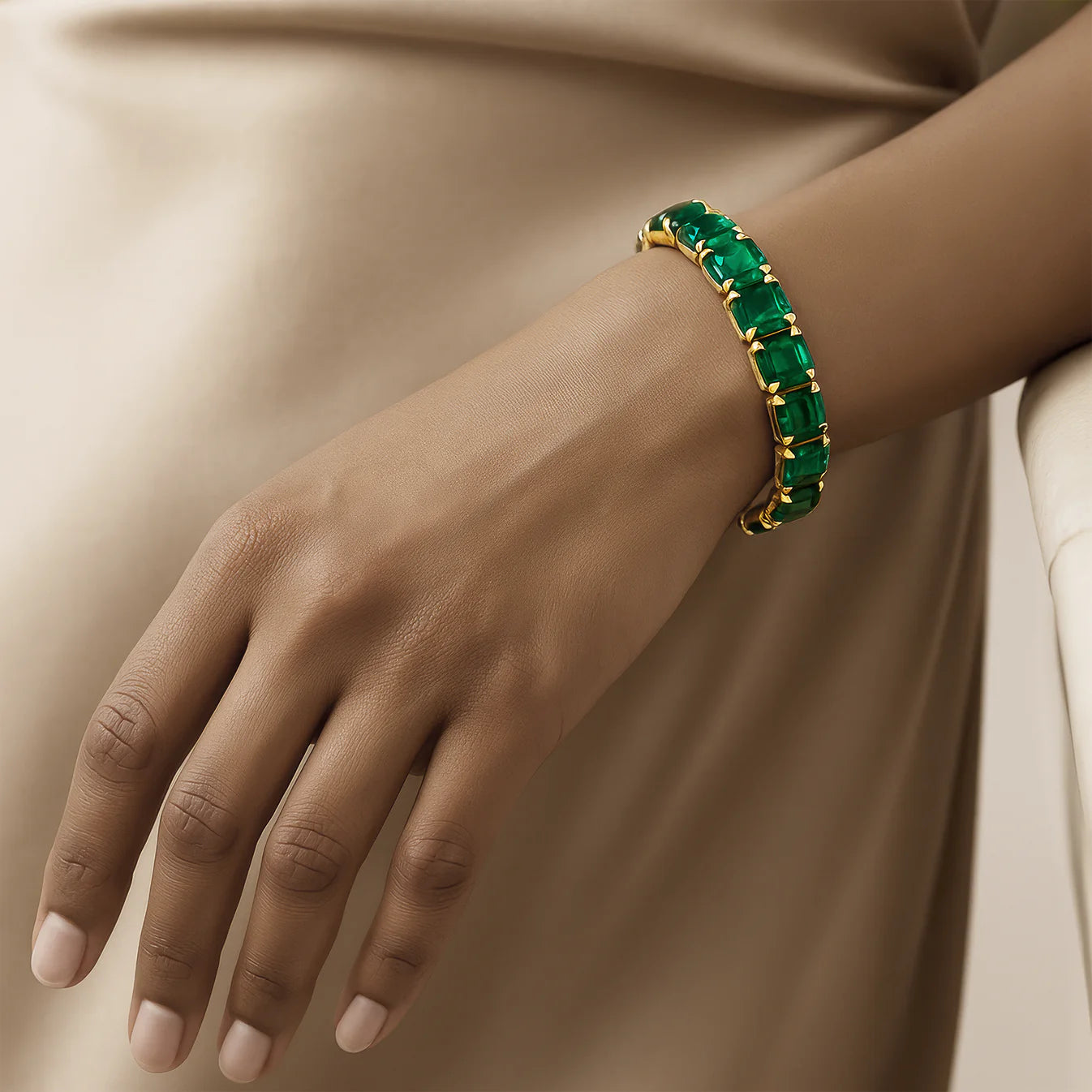 Hand wearing a gold bracelet with green gemstones on a neutral background