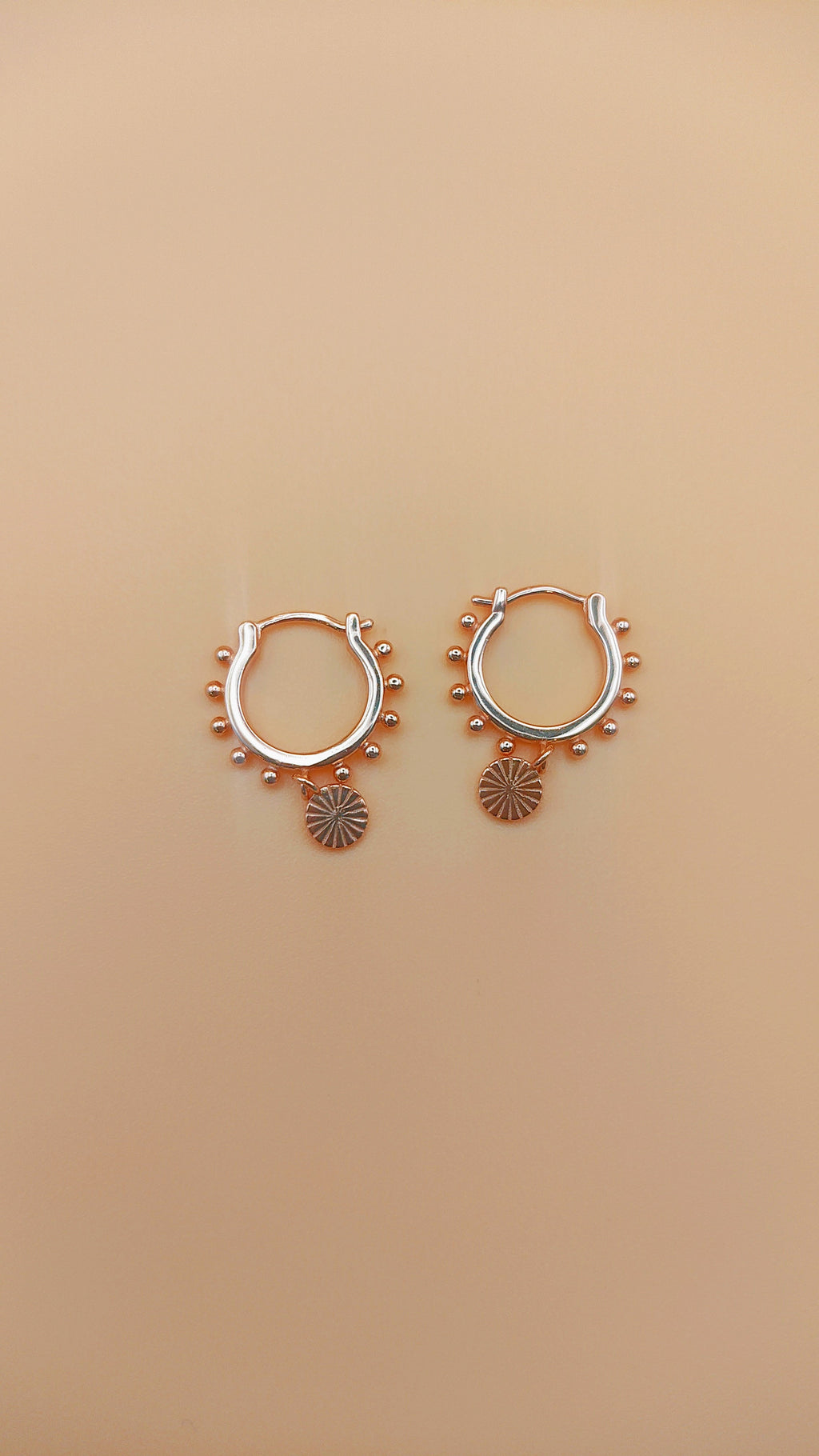 Pair of rose gold hoop earrings with small circular designs on a beige background