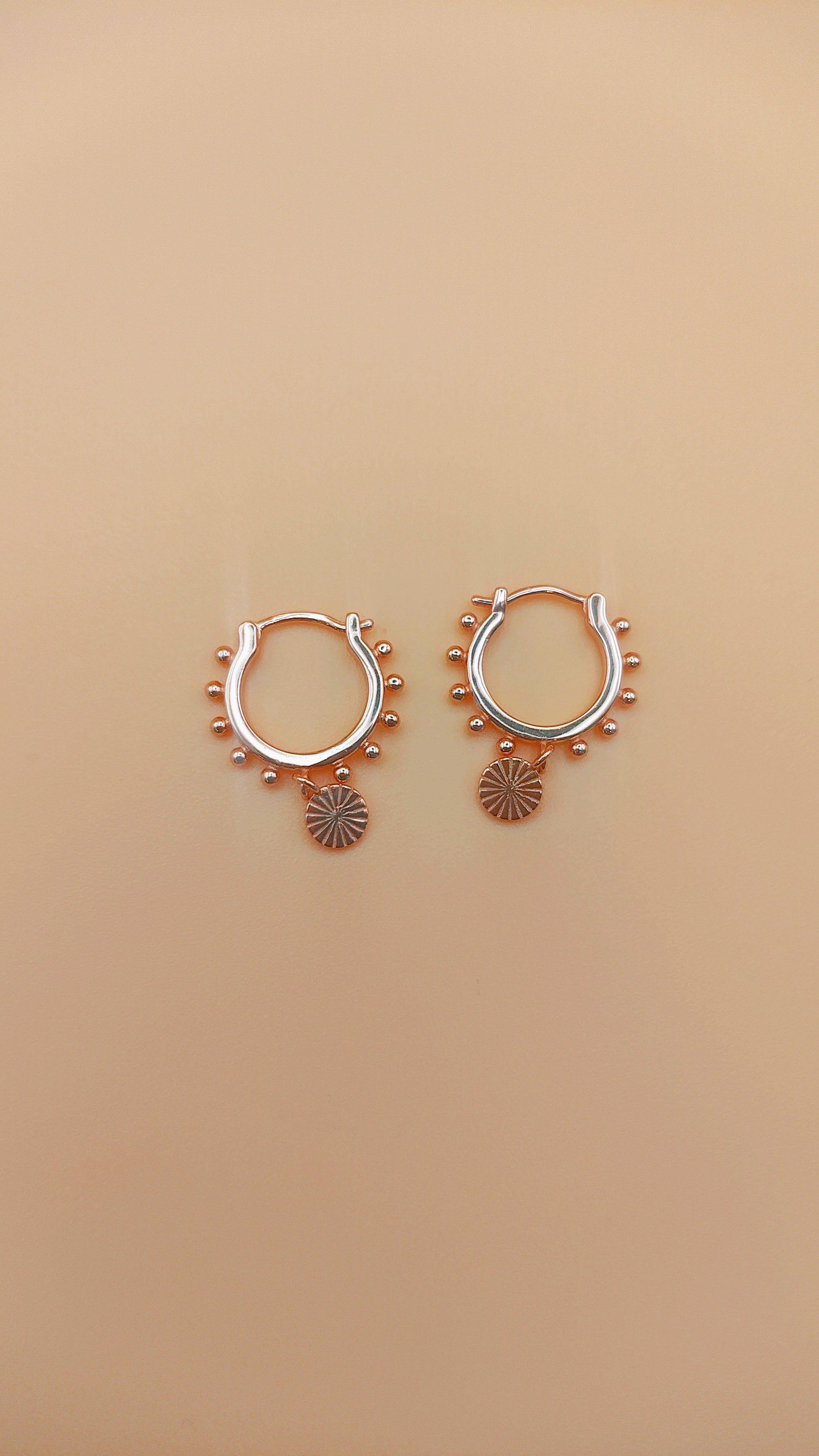 Pair of rose gold hoop earrings with small circular designs on a beige background